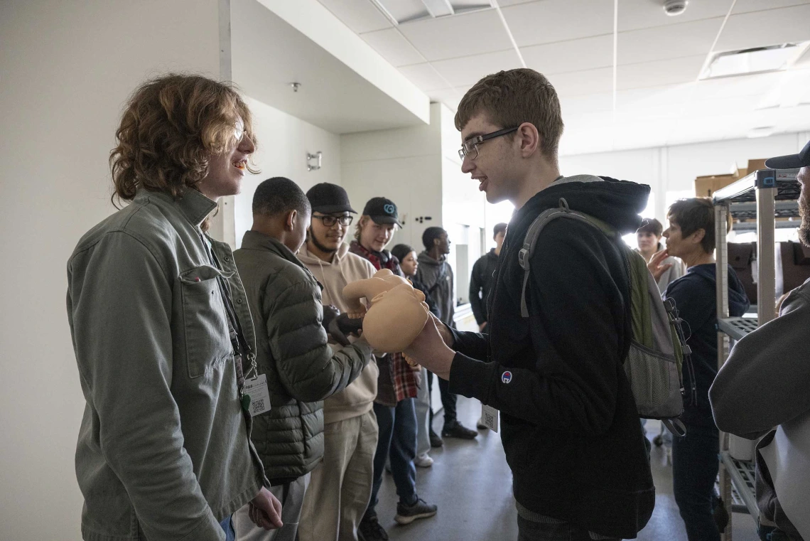 A line of high school students pass a baby doll to each other as they stand in a large room. 