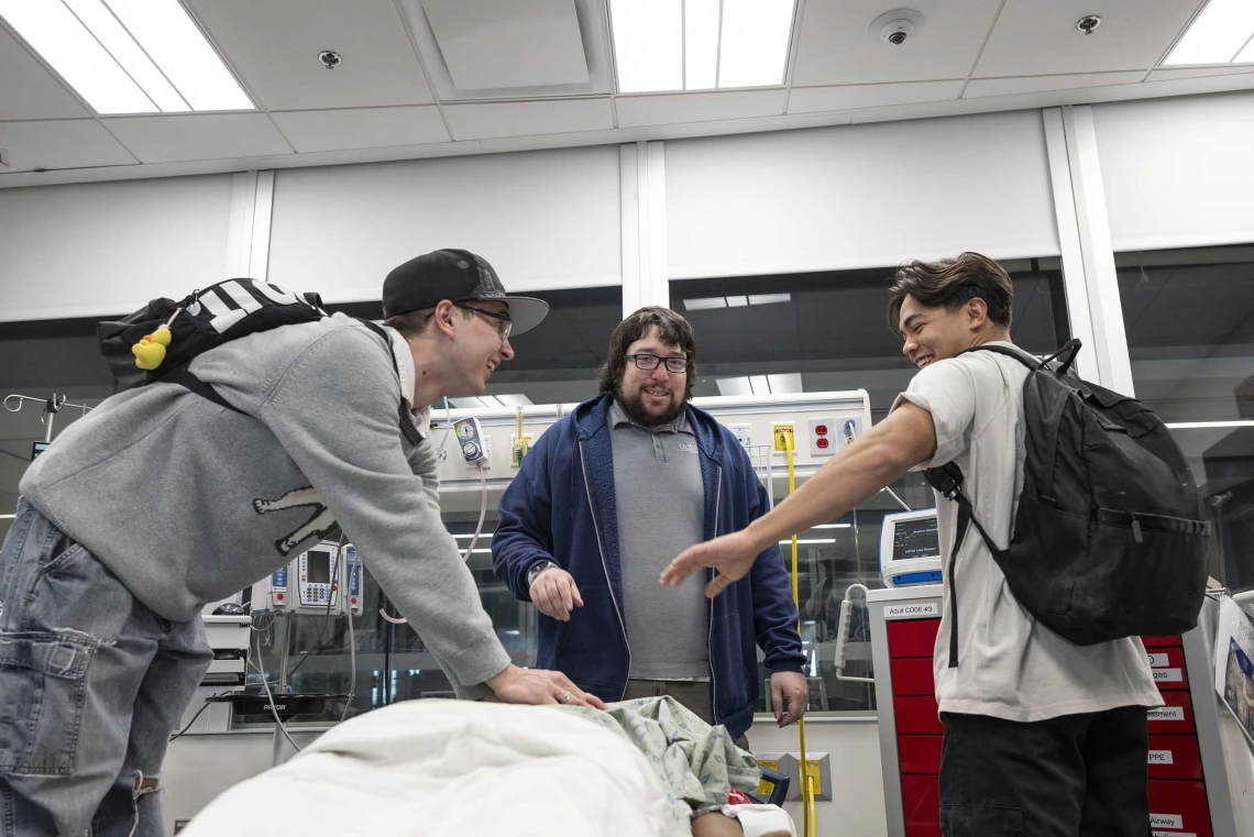 Two high school students laugh as they perform CPR on a manikin while an instructor looks on in a medical training room. 