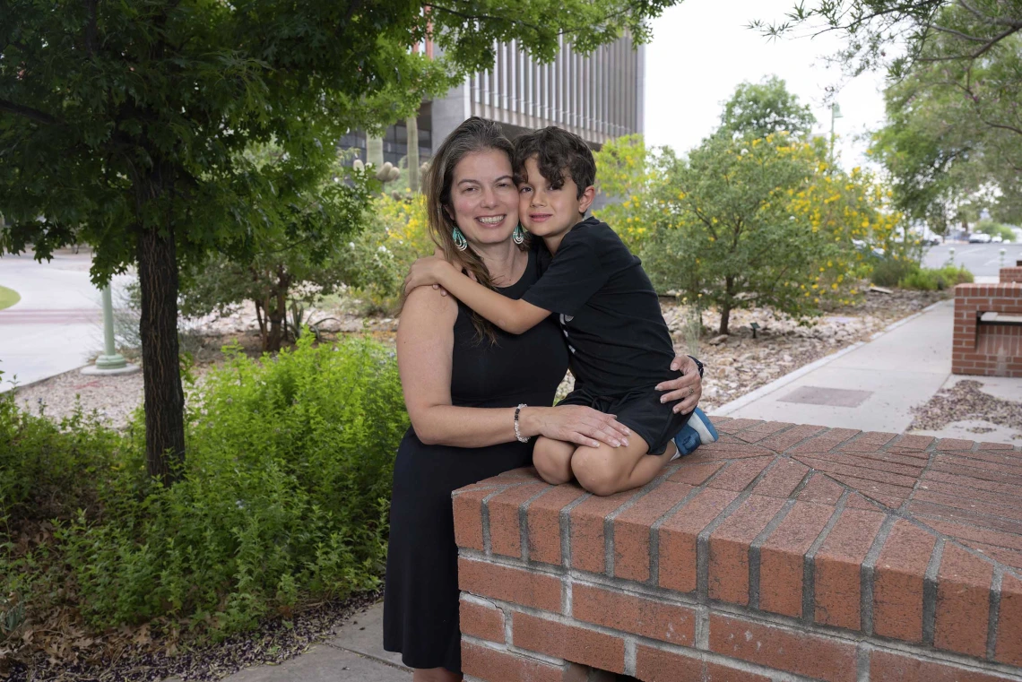 A young boy kneels on a raised surface and reaches out to hug his mother