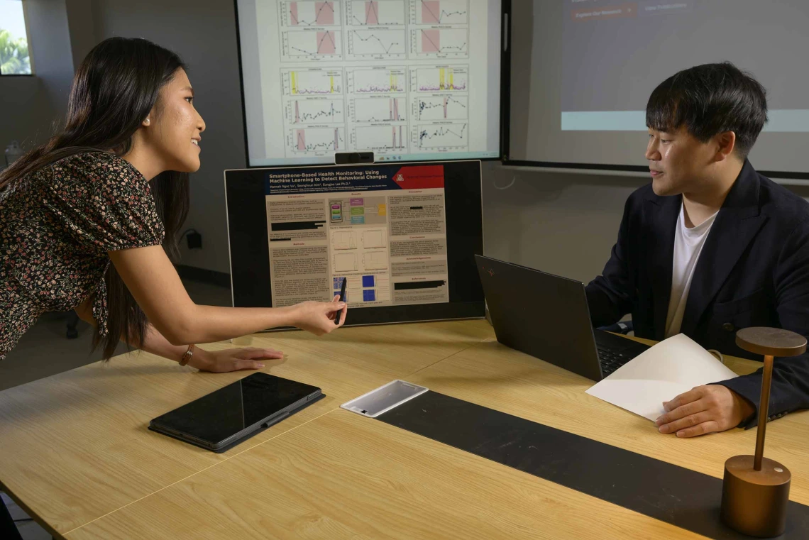 A graduate student, pointing to a small digital monitor, explains a research poster to a faculty mentor
