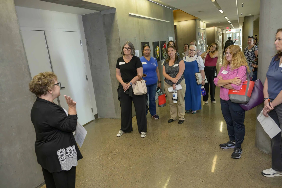 A woman with her back to the camera, one hand raised, speaks to a group of educators gathered outside an elevator at the Phoenix Bioscience Core. 