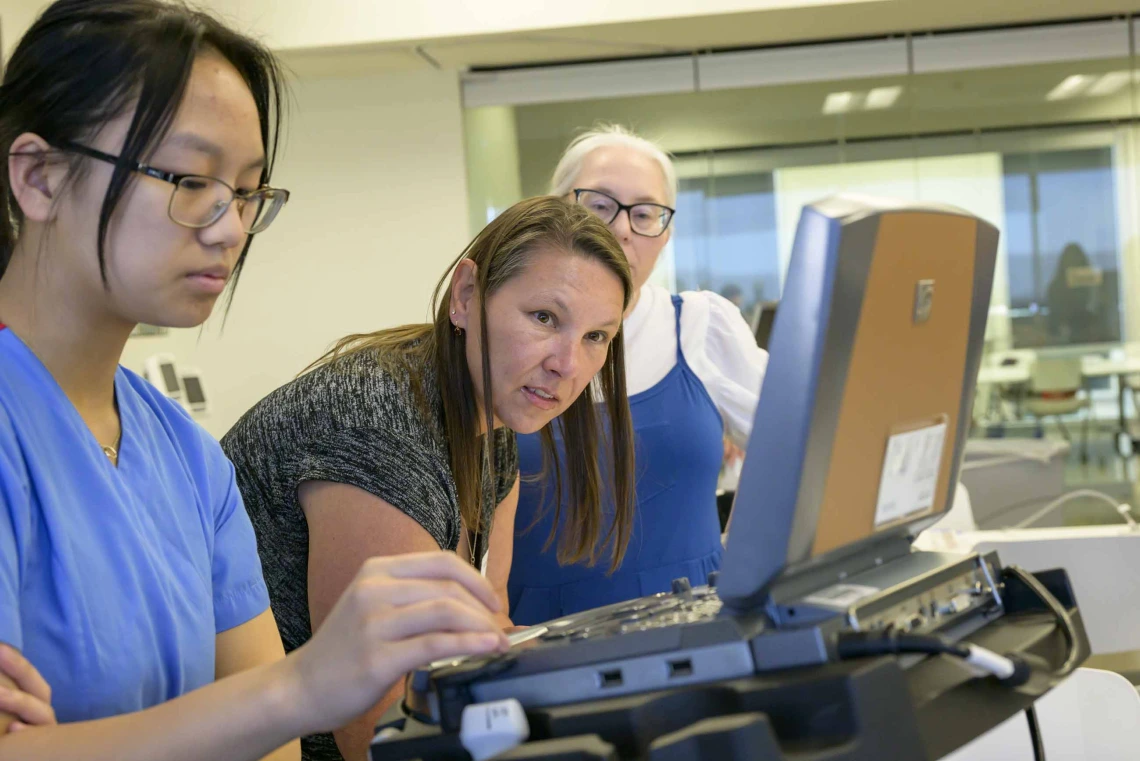 A teacher performing an ultrasound simulation looks at the image, while a fellow teacher watches and a medical student runs the monitor. 