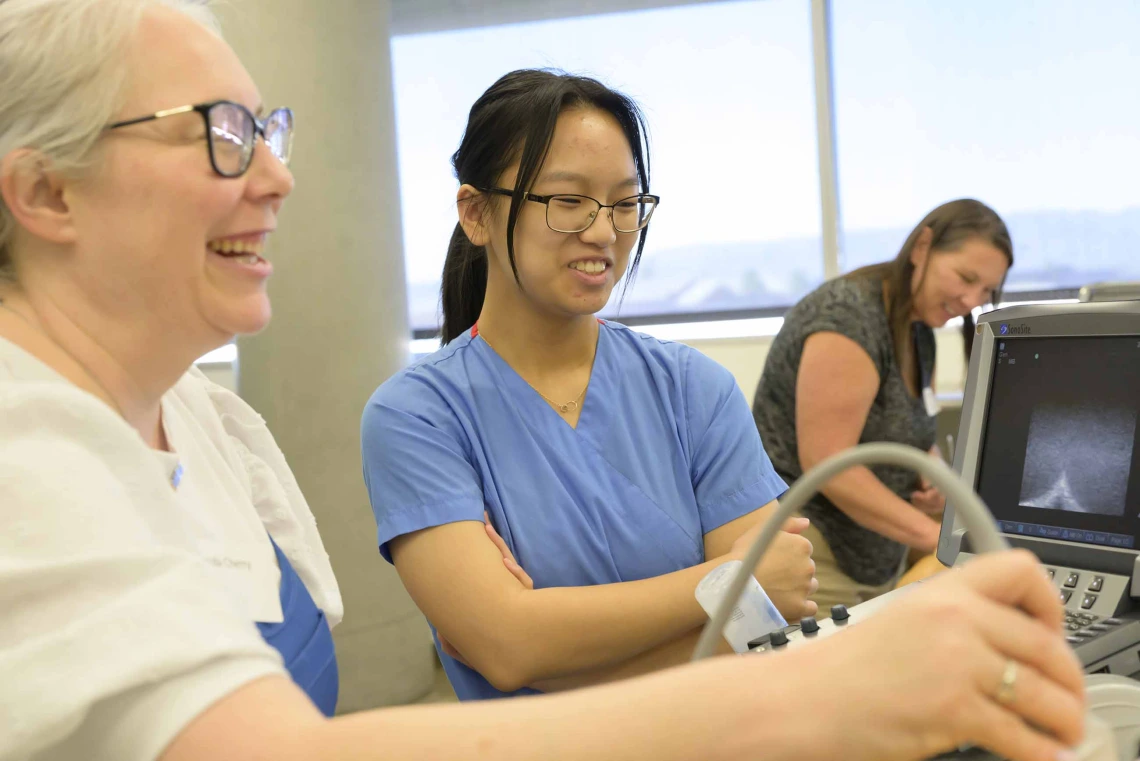 A smiling person uses an ultrasound wand on a manikin as a smiling person in scrubs stands next to her