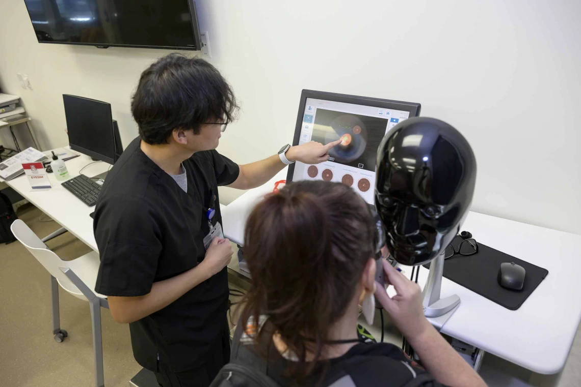 A person with his back to the camera points at a computer screen displaying a virtual eye image while another person peers into an ophthalmology simulator
