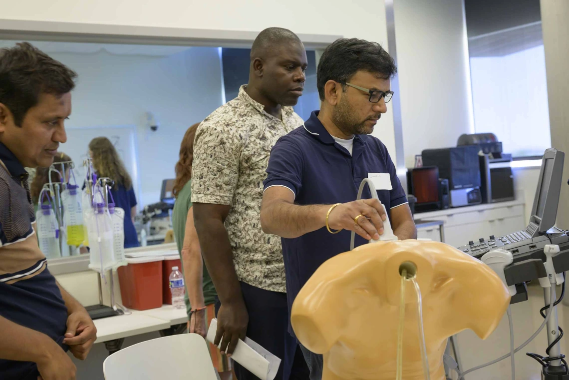 Three educators gather around an ultrasound station where one holds a wand over a manikin, viewing a monitor displaying simulated health conditions, while the others observe. 