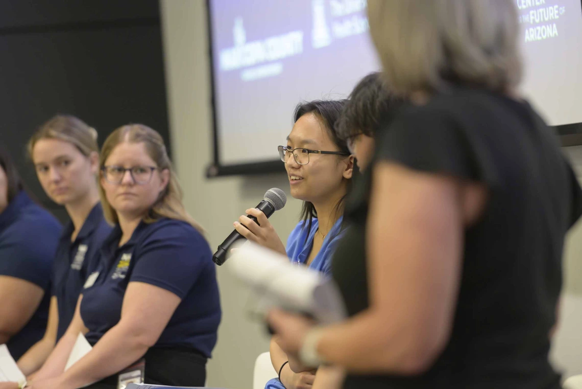 A person holds a microphone, speaking in a conference room, with four people seated on other side of her as part of a panel discussion.