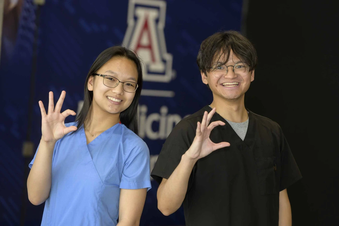 Two people stand side by side, smiling and holding up University of Arizona Wildcat signs with their right hands.