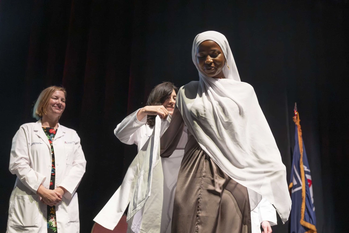 A medical student wearing a hijab is assisted in putting on a medical white coat by a professor in a medical white coat. 