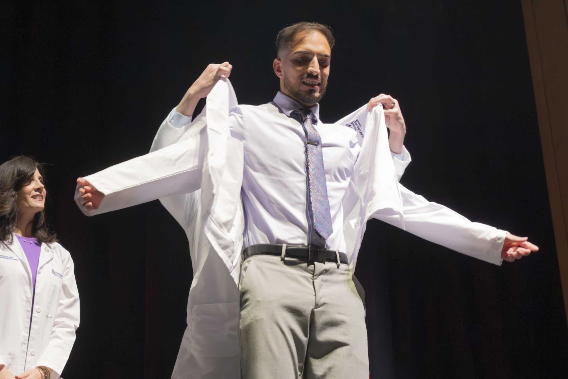 A medical student puts on his white coat with the help of a professor who is standing behind him.