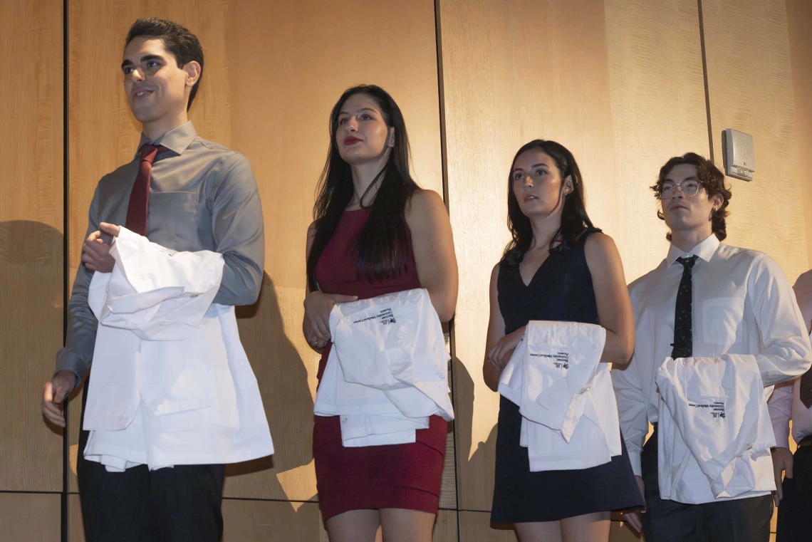 Four University of Arizona College of Medicine – Phoenix students stand in a line with medical white coats draped over their arms. 