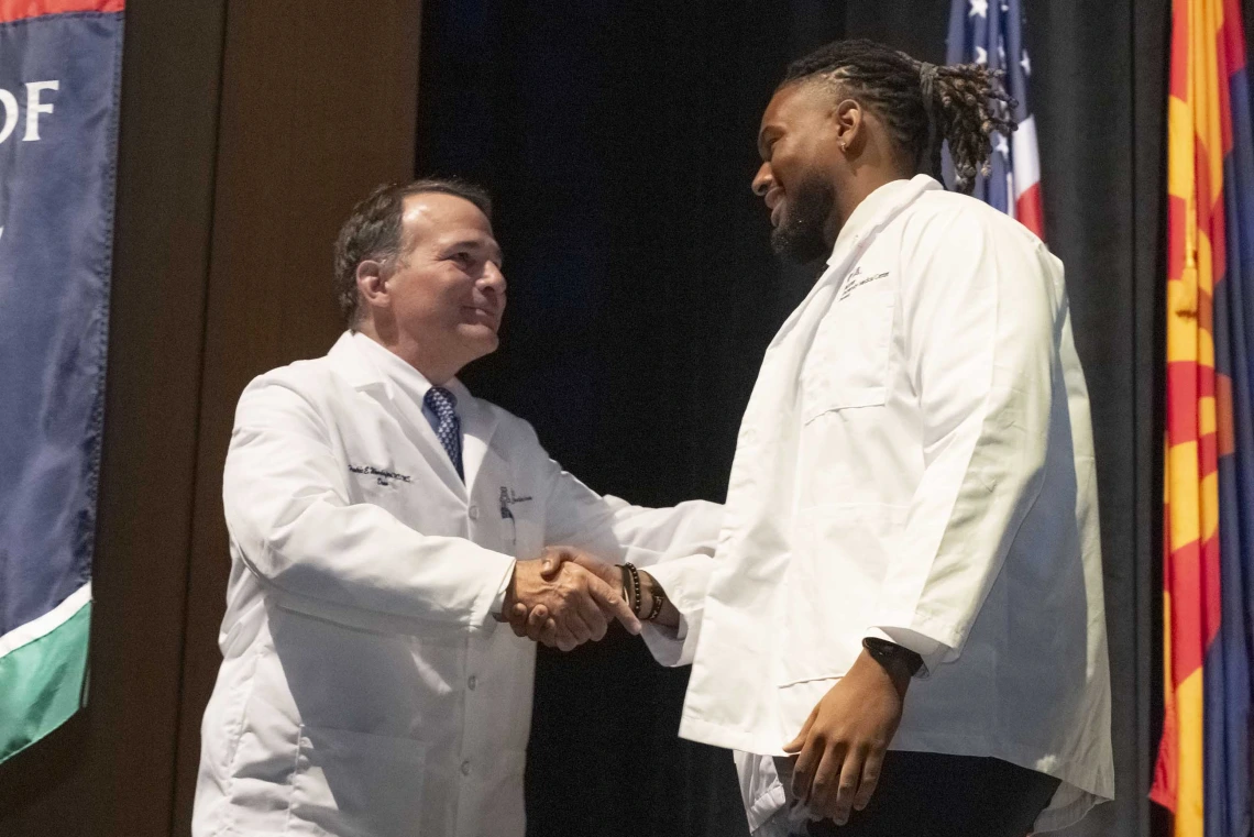 Fred E. Wondisford, MD, MS, MBA, shakes hands with a new medical student onstage. Both are smiling and wearing medical white coats. 
