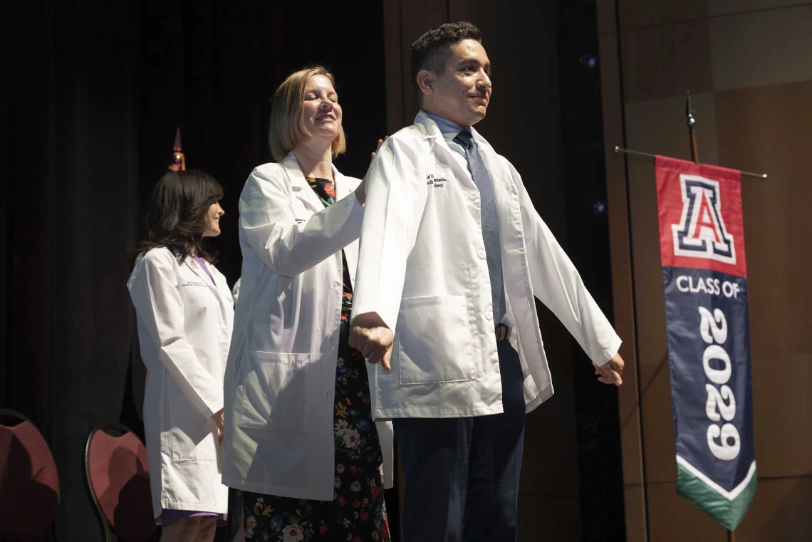 A medical student puts on his white coat with the help of a professor who is standing behind him.