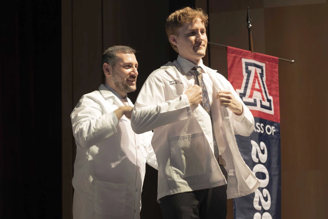 A medical student puts on his white coat with the help of a professor who is standing behind him.