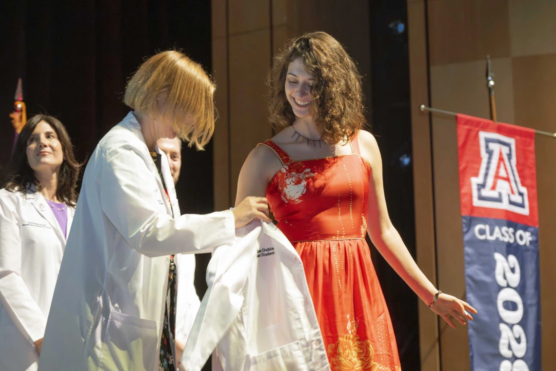 A professor in a medical white coat helps a new medical student put on her white coat during a ceremony. 