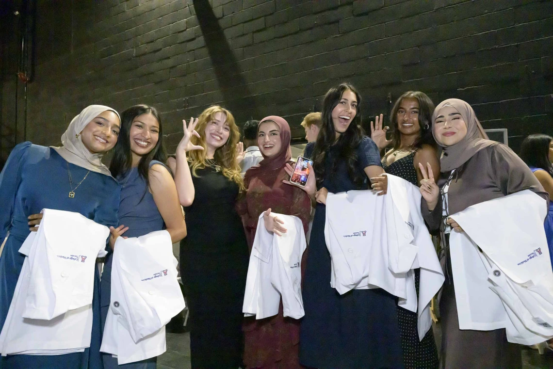 Seven female medical students stand together, smiling, while holding medical white coats over their arms. 