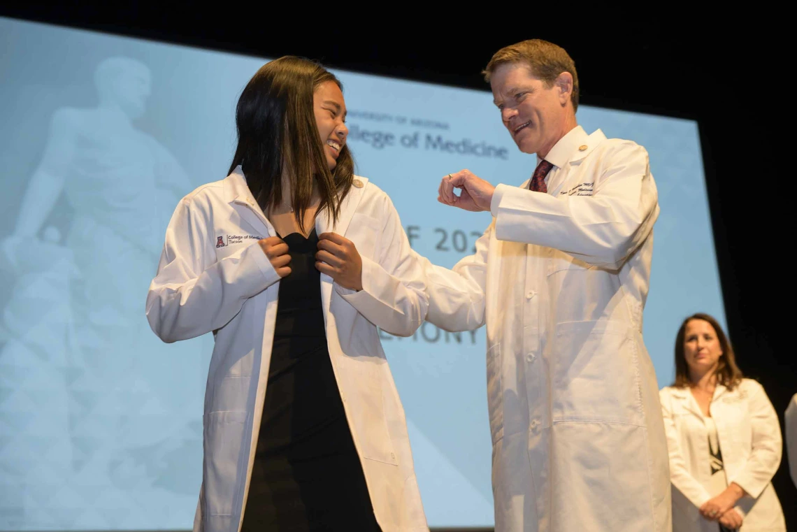 A first-year medical student wearing a medical white coat looks back and smiles at her mentor, a doctor who is also wearing a medical white coat, while standing onstage.  