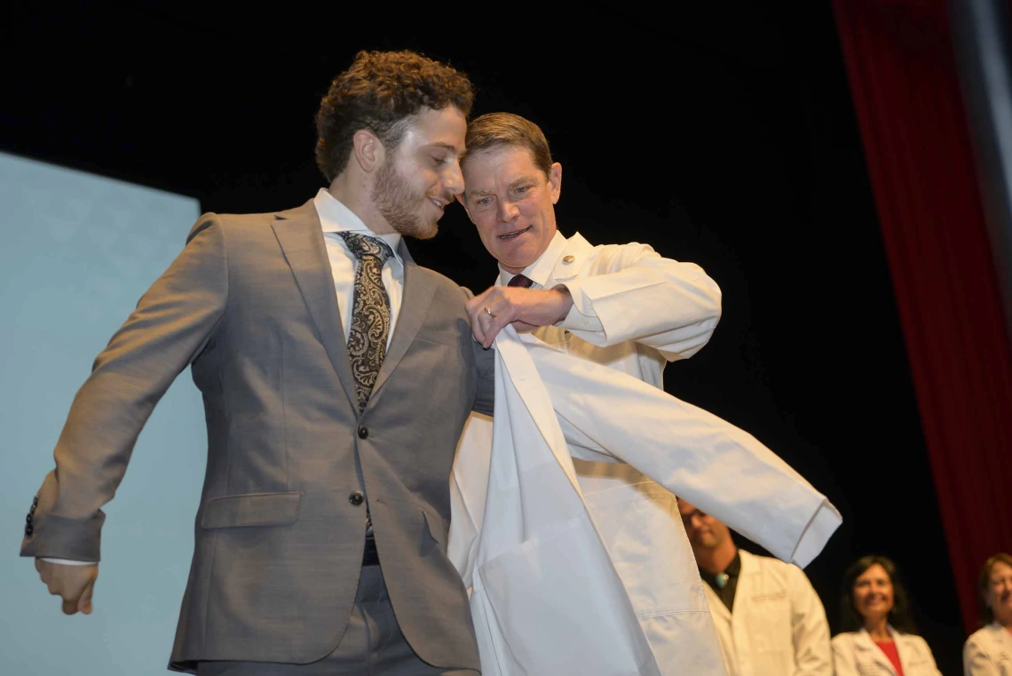 A new medical student smiles as a professor helps him put on his white coat.