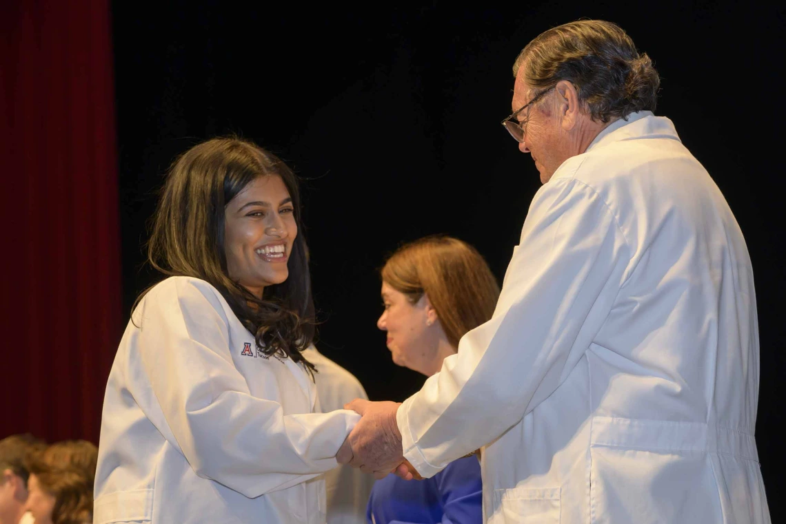 A first-year medical student, smiling and wearing a white medical coat, shakes hands with the dean of the college, who is also wearing a white coat. 