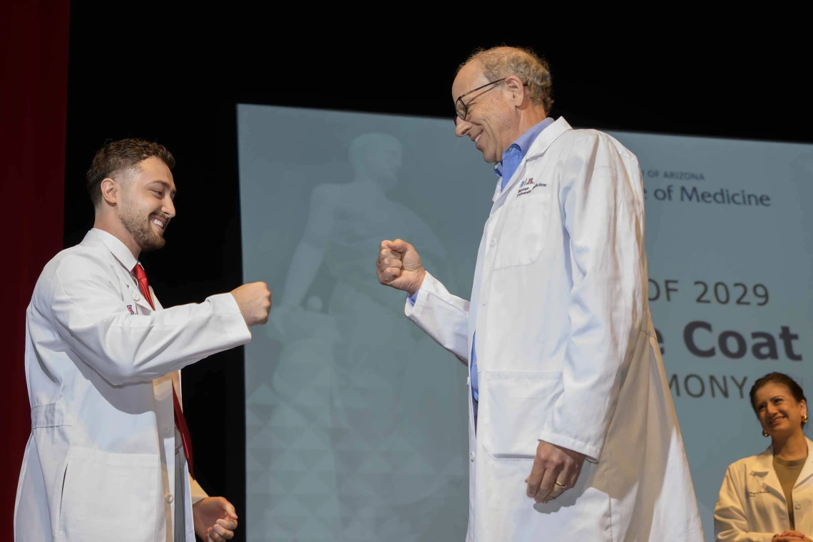 A medical student and his professor, both wearing white coats, smile as they bump fists onstage. 