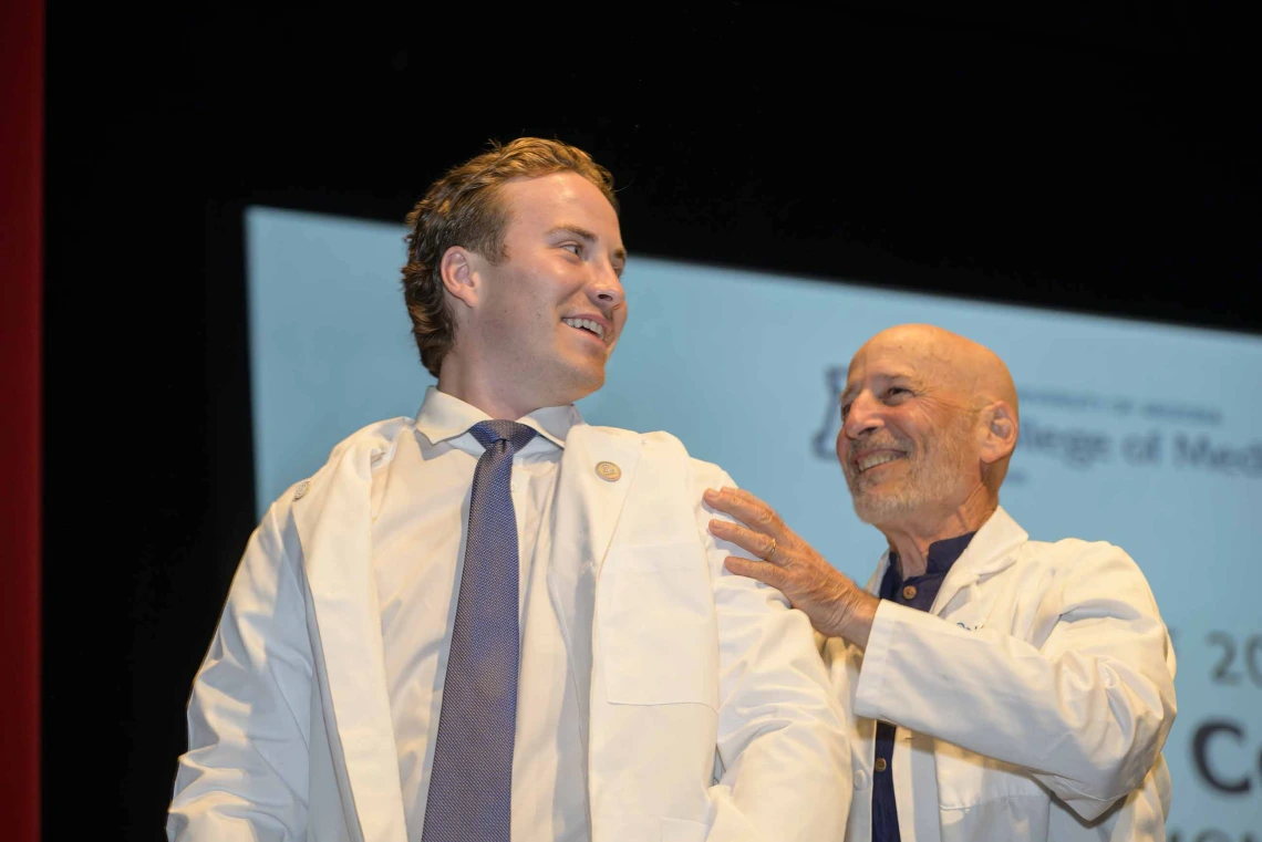 A first-year medical student wearing a medical white coat looks back and smiles at his mentor, a doctor who is also wearing a medical white coat, while standing onstage.  