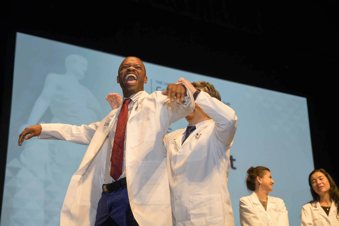 A first-year medical student holds his arms out and laughs as a professor standing behind him helps him put on a medical white coat. 