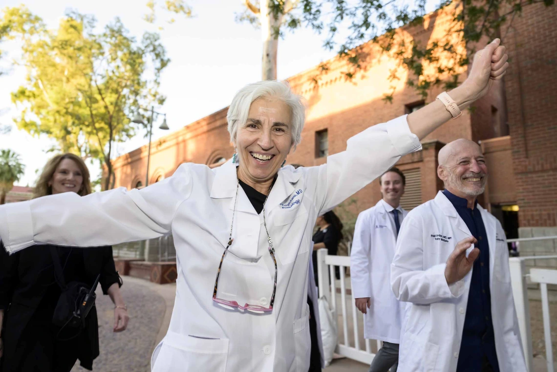 A medical professor in a white coat smiles and raises her arms in celebration while walking with other professors and students outside. 