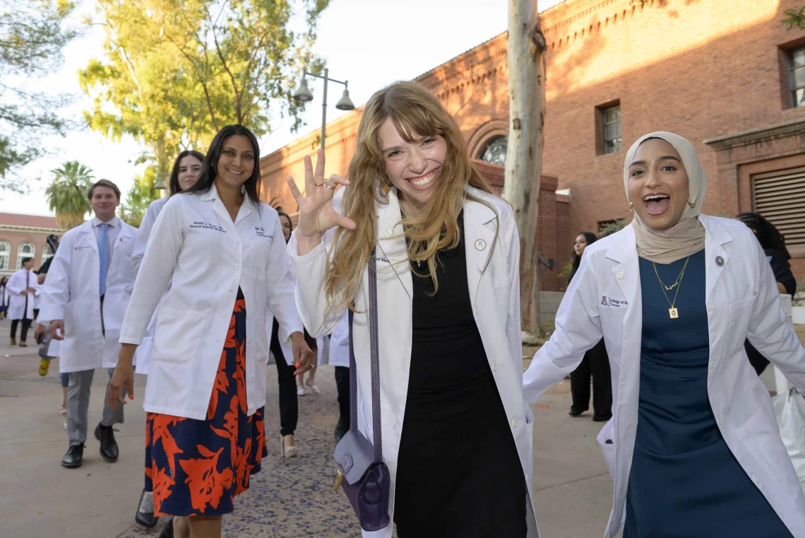 Several first-year medical students wearing white coats smile and celebrate as they walk outside Centennial Hall after their white coat ceremony. 