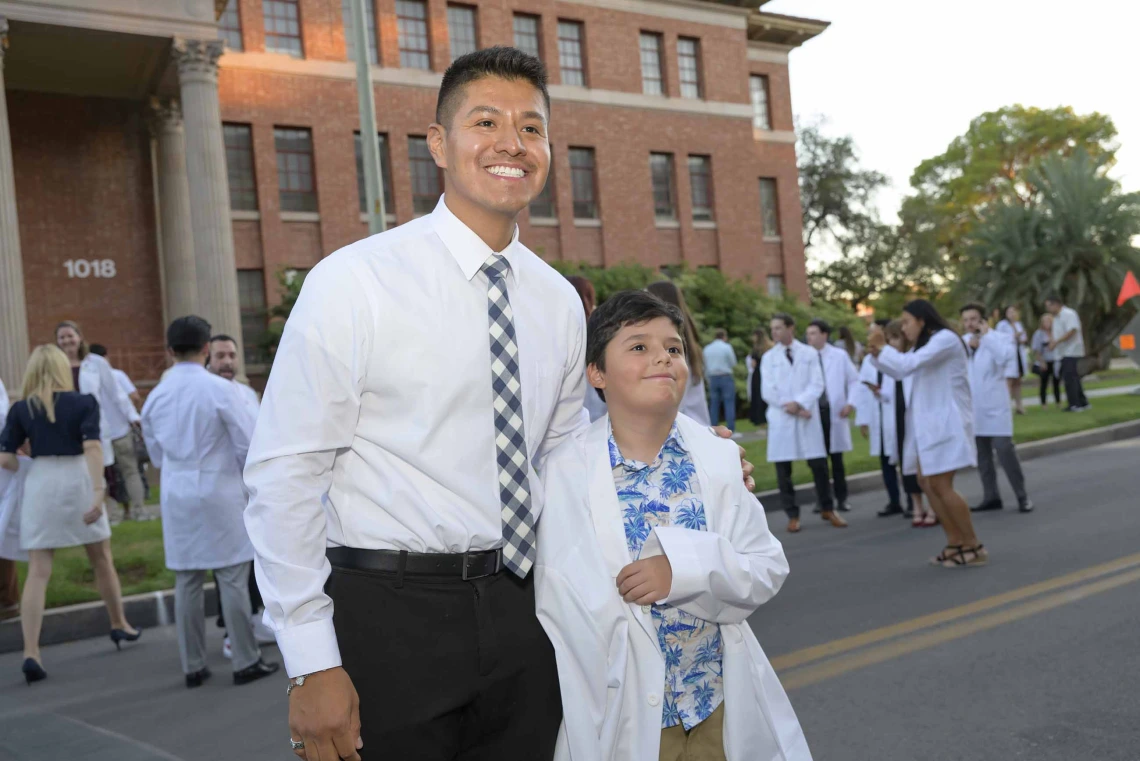 A first-year medical student wearing a shirt and tie stands with his arm around his younger cousin, who is wearing his medical white coat. The coat is very large on the younger cousin. 