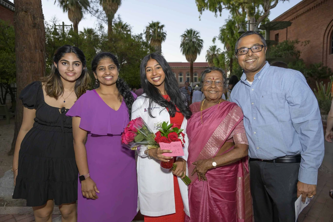 A first-year medical student wearing a white medical coat smiles and holds flowers while standing with smiling family members outside. 