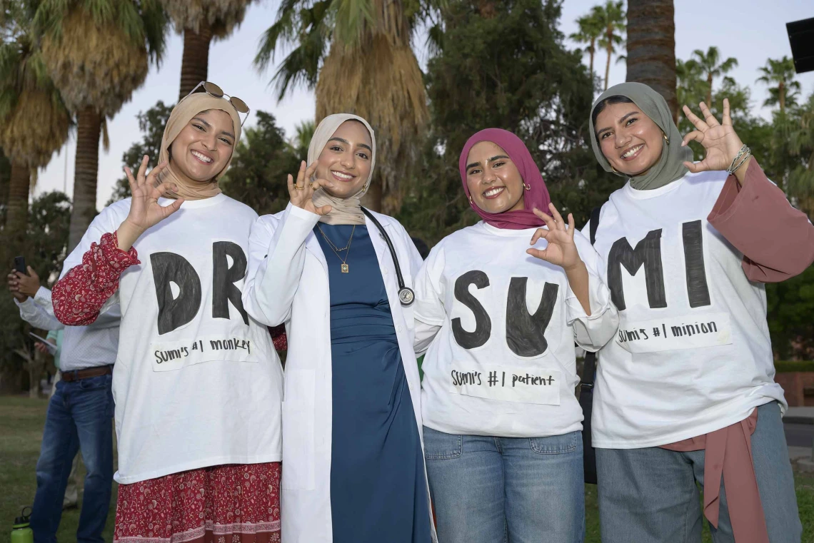 Four smiling young women stand together and flash the U of A Wildcat hand sign. One is wearing a medical white coat and the other three are wearing white T-shirts with large black lettering spelling out “Dr. Sumi,” with two large letters on each shirt. 