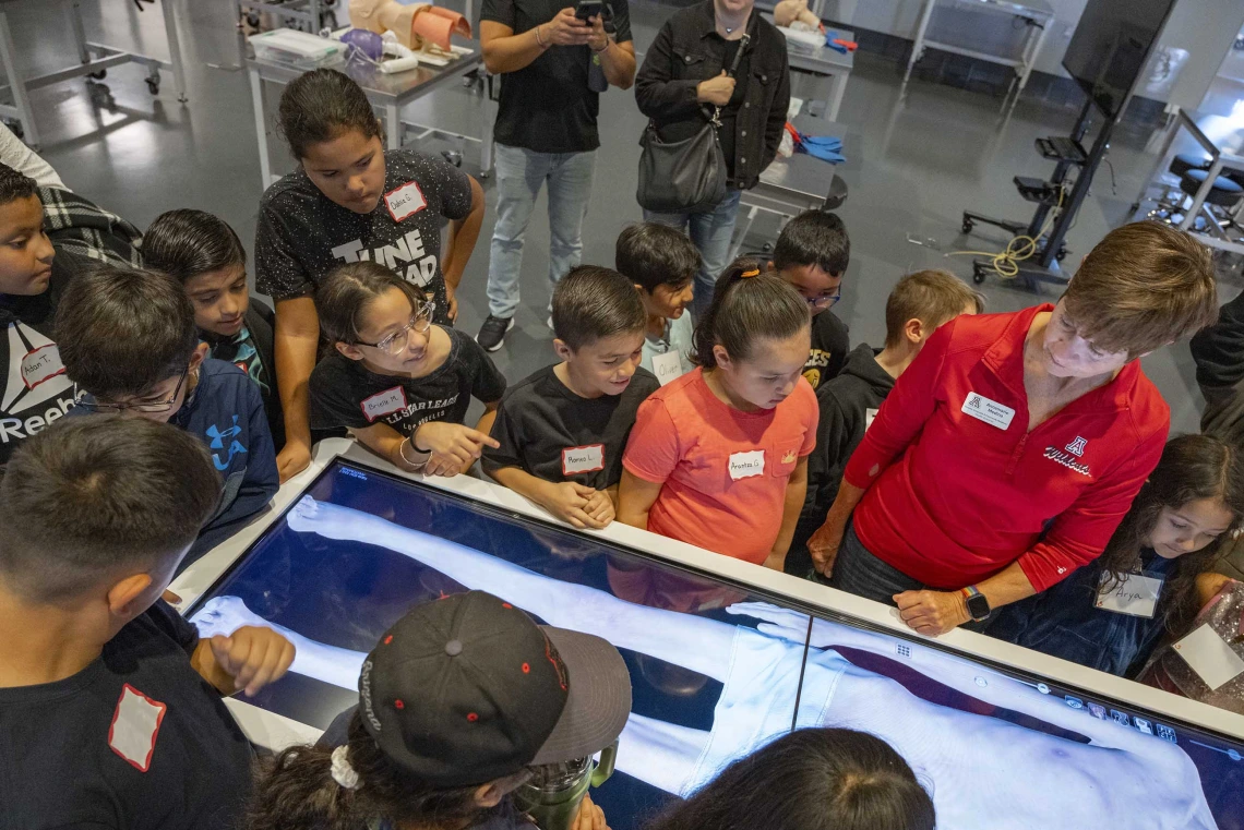 About a dozen grade-school students stand around a table with a digital image of a body while a tour guide demonstrates how it works.