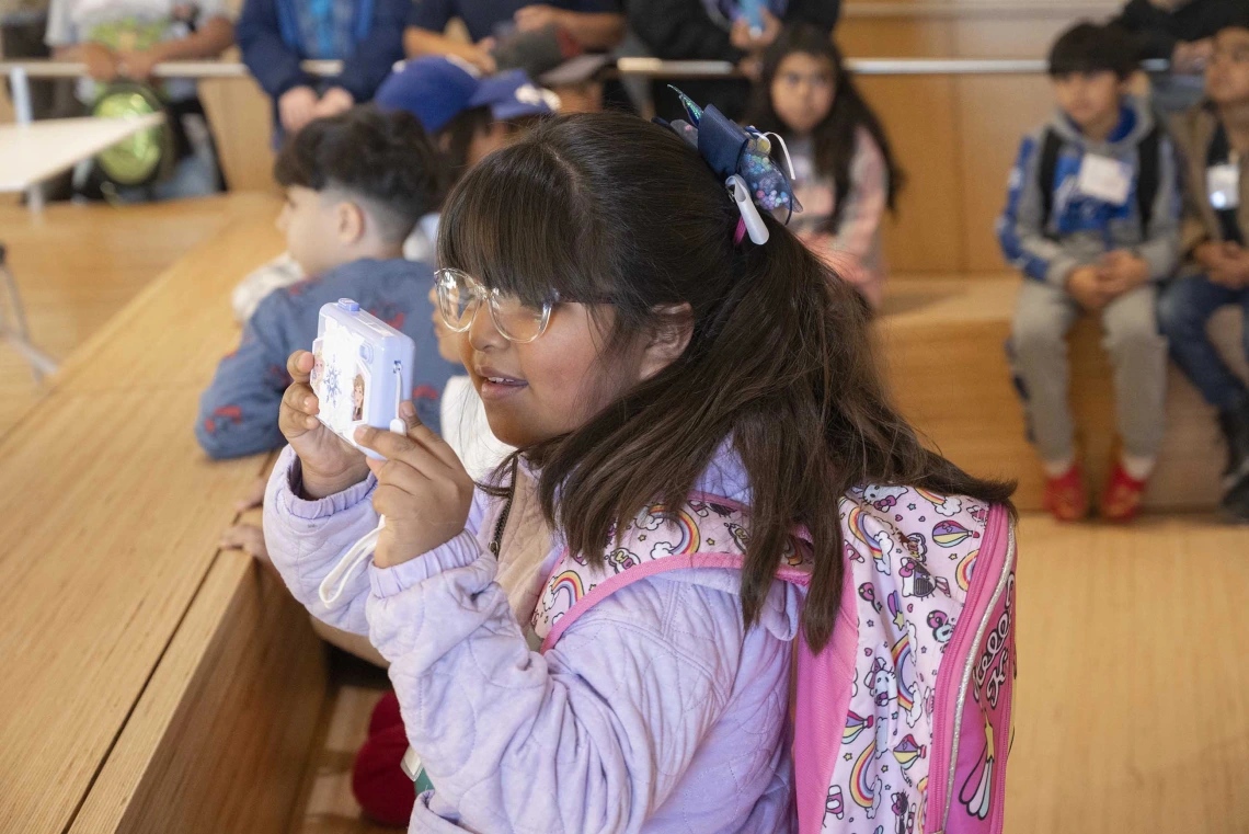 A young girl stands in front of other students while holding up a camera to take a photo. 