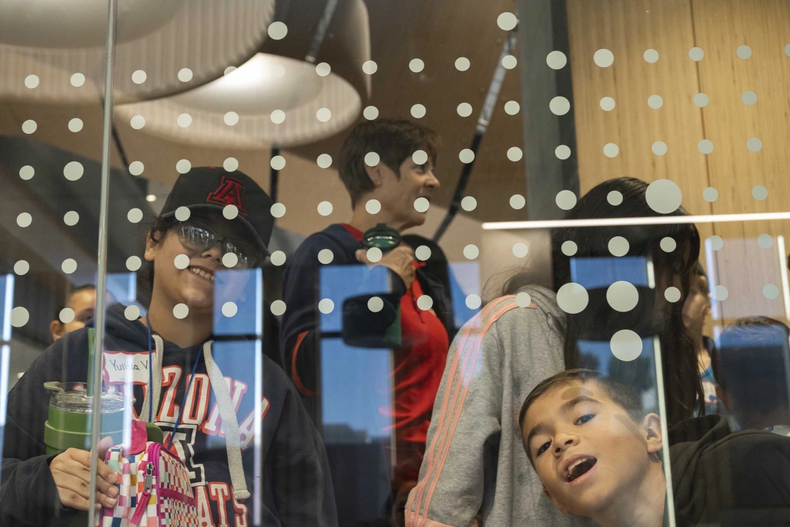 Two young boys make faces through a glass wall as their class walks through a building. 
