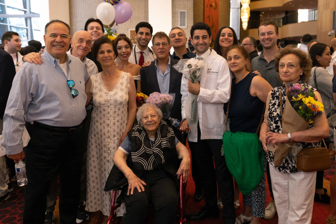 A large family stands around two medical student wearing a white coats in a crowded lobby. 
