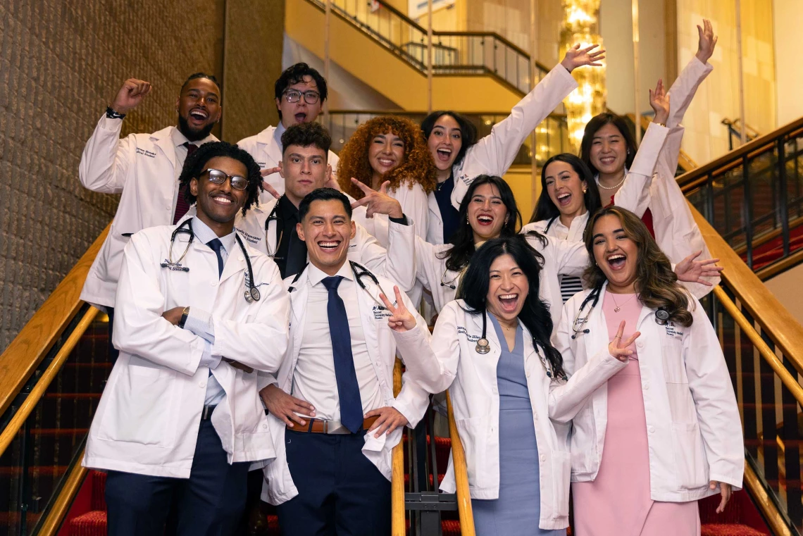 About a dozen students wearing medical white coats stand on the stairs in a lobby, smiling and cheering. 