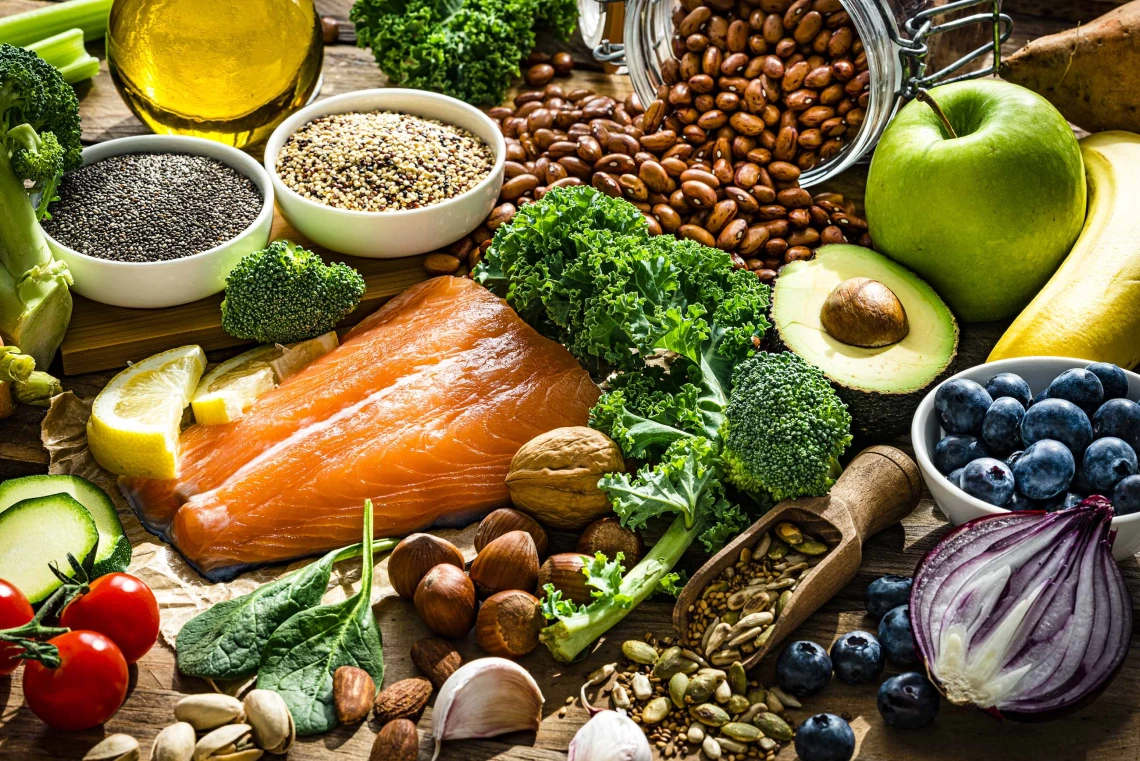 Healthy foods on a table: salmon, kale, spinach, avocado, almonds, blueberries, onion, chia seeds, cucumber, tomato, apple, pistachios, garlic, carafe of olive oil, lemon, broccoli