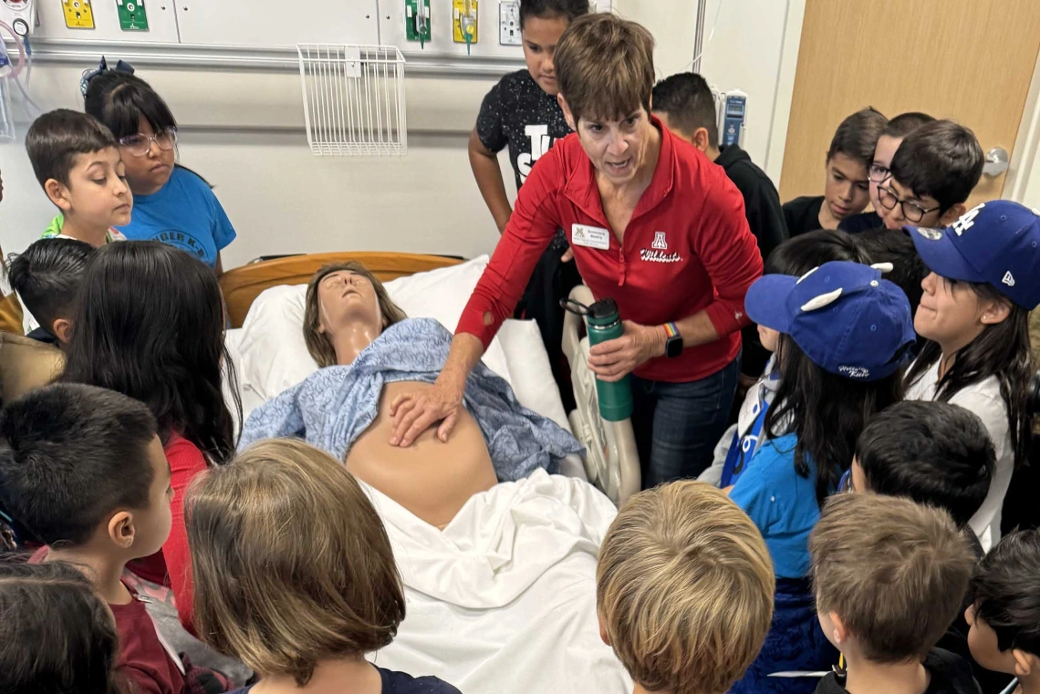 More than 20 elementary school children stand around a pregnant manikin lying on a hospital bed as an adult pushes on the manikin’s abdomen. 