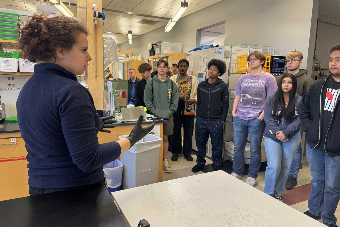 A woman speaks to a group of high school students in a science lab.  