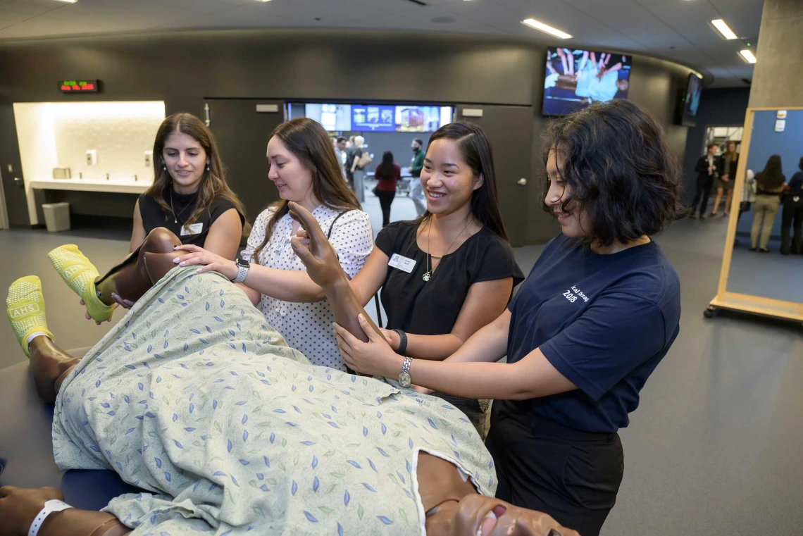 Several students stand alongside a simulation manikin dressed in a hospital gown. 