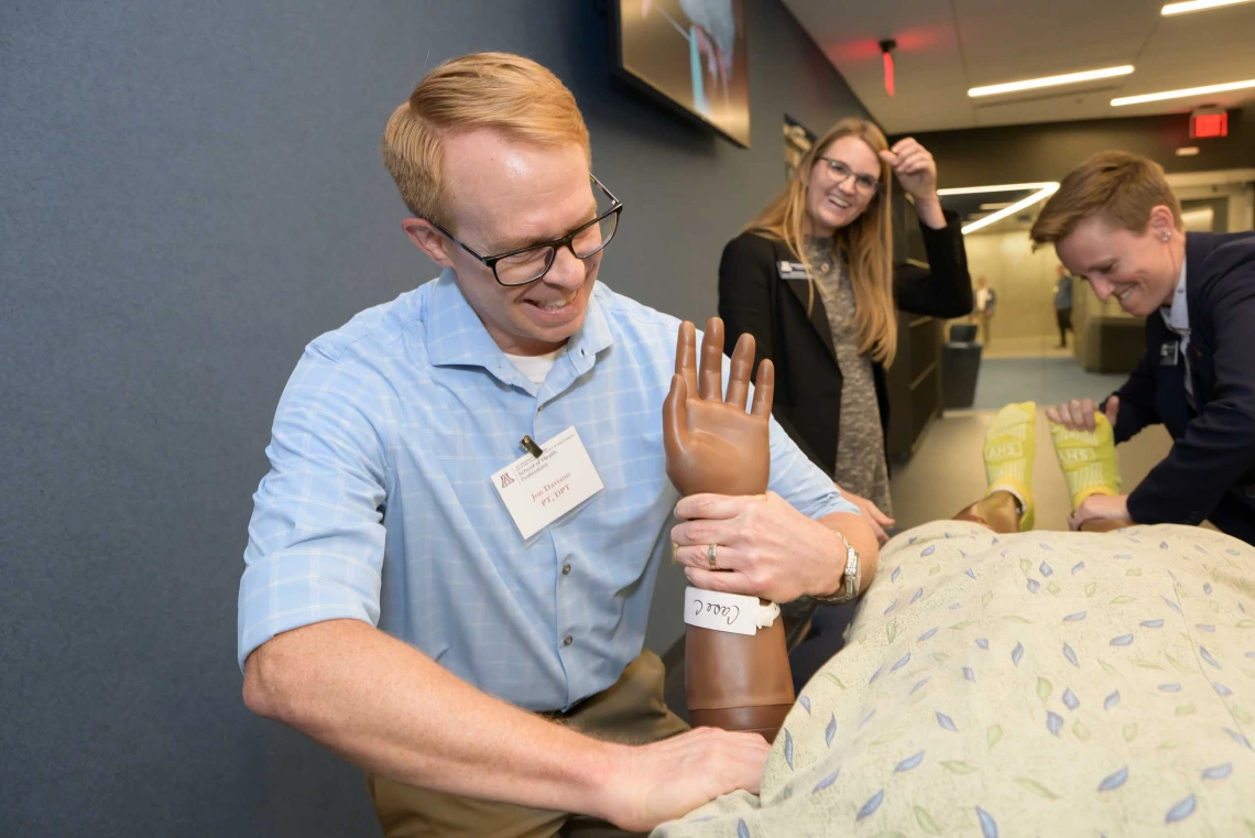 A man grasps the wrist of a simulation manikin dressed in a hospital gown.