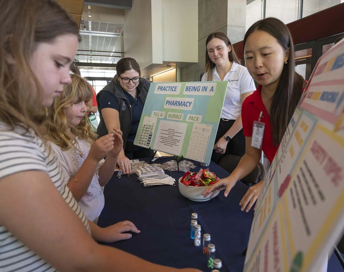 Two young girls look at a display that reads “Practice Being in Pharmacy” as three young women standing behind posterboards point things out to them.