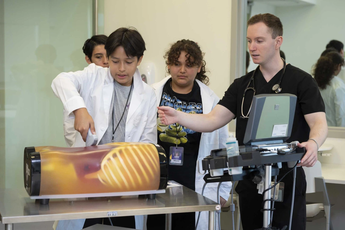 A 13-year-old boy holds a medical tool against a model of a torso while a medical student in scrubs and two other middle schoolers watch.