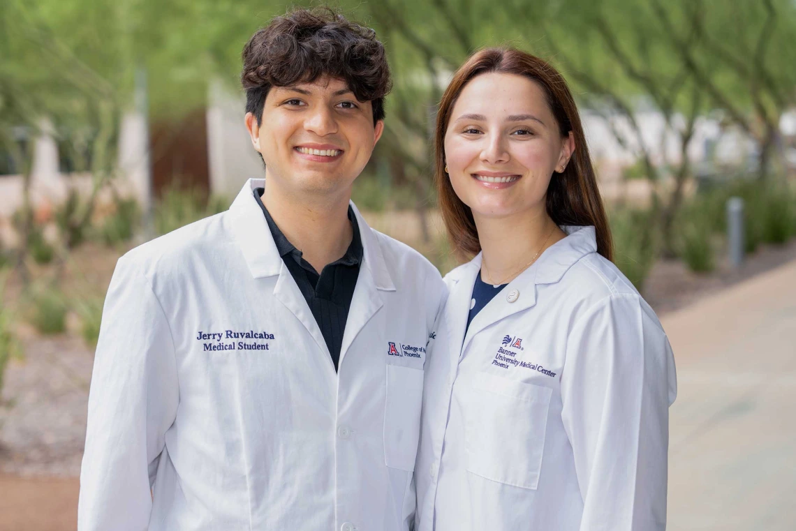 Medical students Jerry Ruvalcaba and Katie Simic stand side by side outdoors, smiling in their white lab coats embroidered with titles from the University of Arizona College of Medicine - Phoenix.