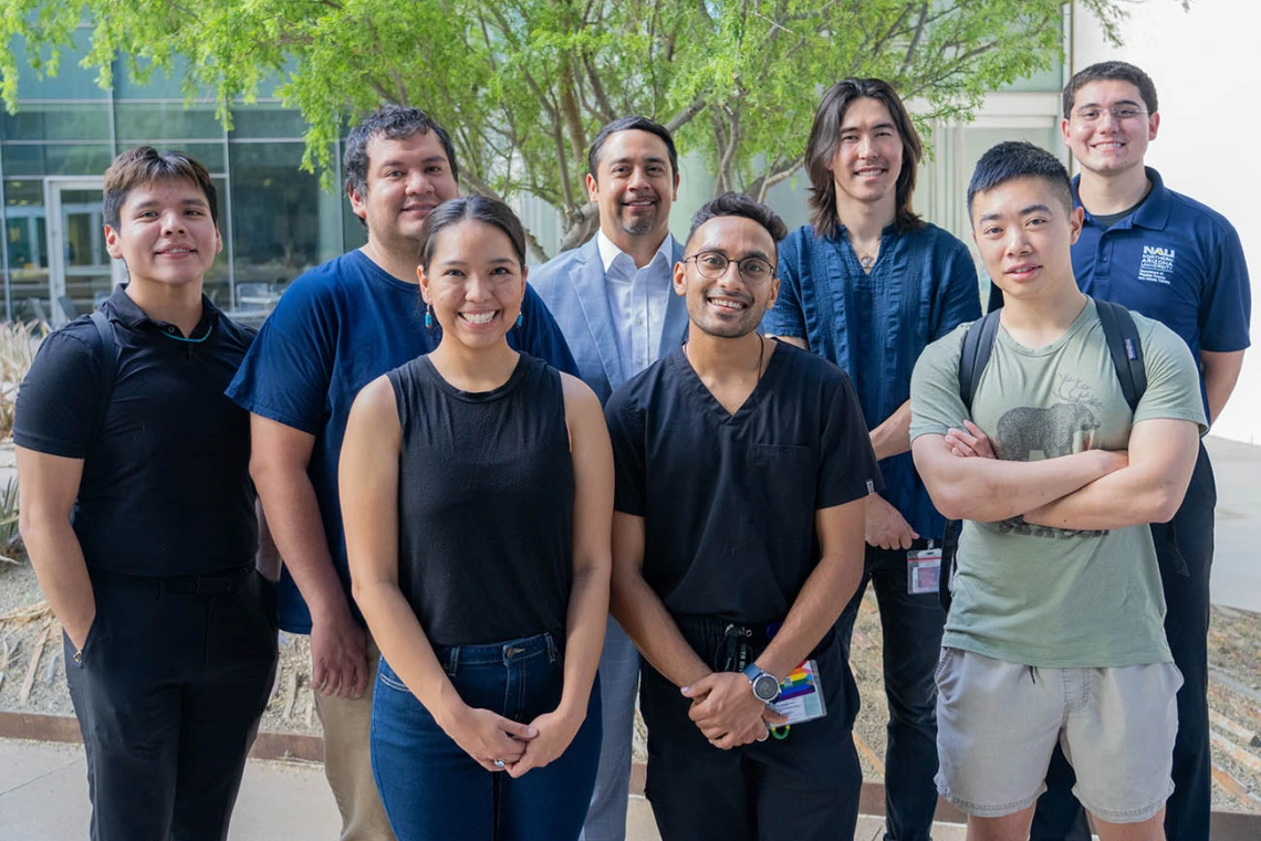 Eight people stand in a group in an outside setting all smiling. 