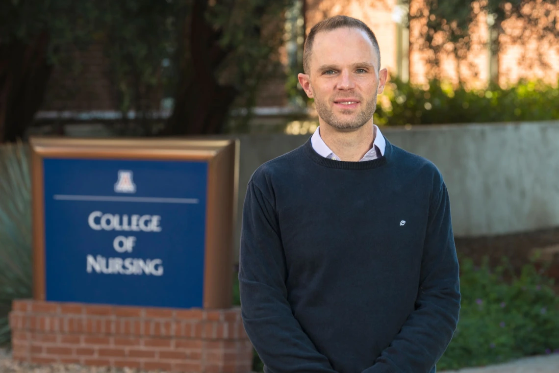 Man smiling slightly and wearing a sweater outside. Behind him is the University of Arizona College of Nursing sign