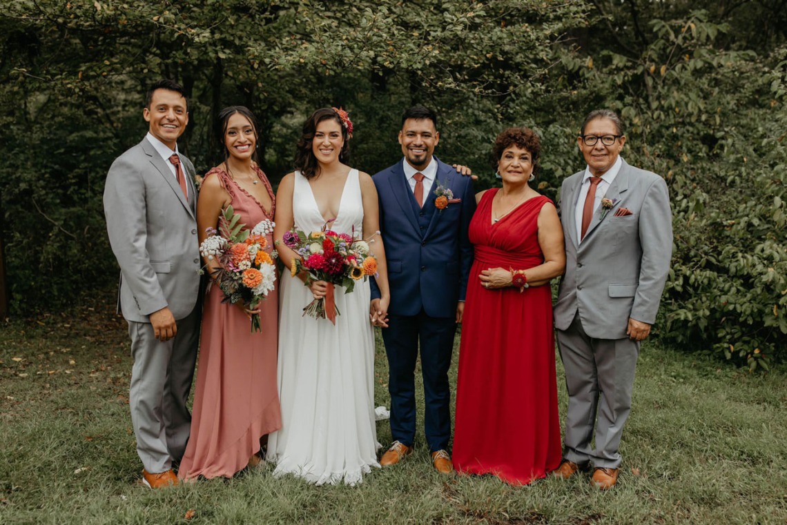 A bride and groom smile surrounded by family members in an outdoor setting