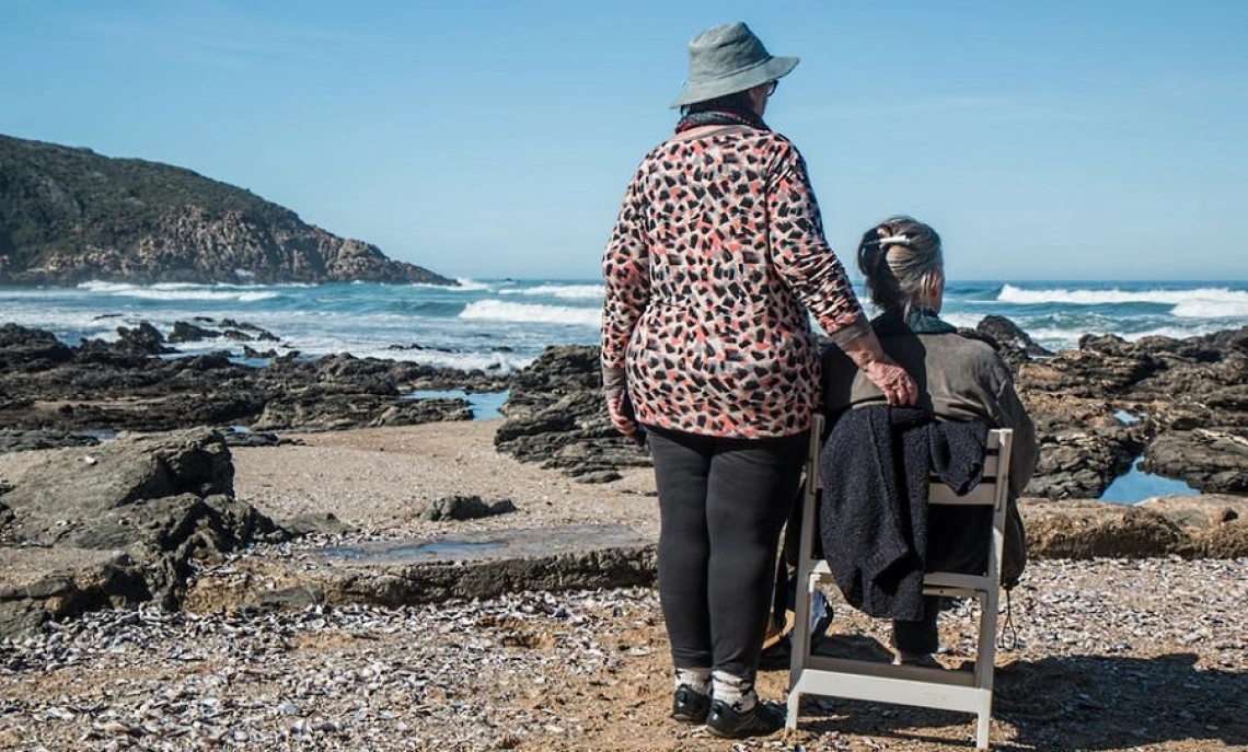 Caregiver standing next to a woman in a white chair in front of body of water