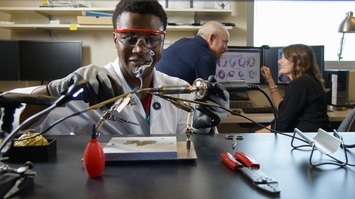 Ike Chinyere solders wiring for a cardiac electrophysiology model to study heart arrhythmias. Steven Goldman, MD, and Elizabeth Juneman, MD, research mentors, in background.