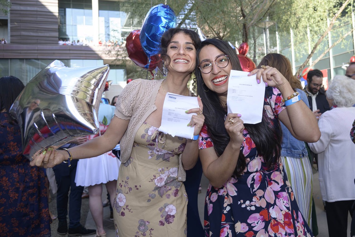 Two women in dresses with long dark hair hold up match day letters and smile big