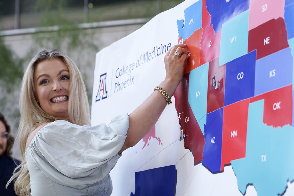 A light skinned woman with blonde hair smiles big as she puts a pin in a big map of the U.S. 
